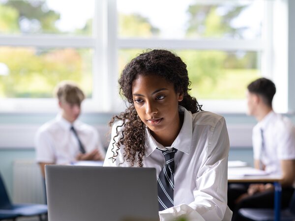 A secondary student in school uniform concentrating on a laptop in a bright classroom, two classmates working behind her