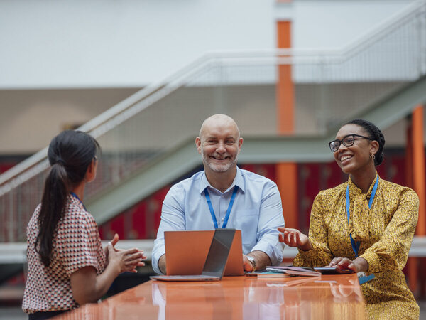 Three members of school staff talking around a laptop at a table in a school atrium, lanyards on, one explaining and the others listening